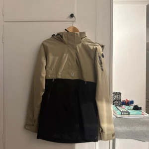 Beige och svart skidjacka 8848 Altitude - Snygg beige och svart skidjacka från 8848 Altitude med huva, dragkedjor och praktiska fickor. Jackan har en sportig look med logga på bröstet och är perfekt för dig som gillar stilren design. Materialet är slitstarkt och vattenavvisande syntet.