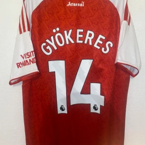 Arsenal Gyökeres 14 matchtröja Adidas S - Snygg Arsenal fotbollströja från Adidas med Gyökeres och nummer 14 på ryggen. Röd bas med vita detaljer, klubbmärke och sponsortryck framtill. Kortärmad modell i lätt och ventilerande material, perfekt för match eller träning.