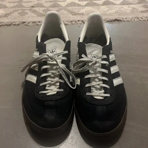 Adidas Handball Spezial svart/vit - Säljer ett par Adidas Handball Spezial sneakers i svart och vitt med klassiska tre ränder på sidan. Ovandelen är i mocka och skinn med vita snören och detaljer. Snygg retrostil och gummisula som ger grymt grepp. Perfekta för dig som gillar streetwear och sportig vibe.