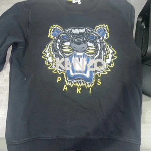 Svart Kenzo sweatshirt med tigertryck - Svart sweatshirt från Kenzo med ikoniskt broderat tigerhuvud i blått, gult och vitt framtill. Tröjan har rund hals, ribbade muddar och är tillverkad i mjuk bomull. Perfekt statement-plagg för dig som gillar streetstyle och vill sticka ut.