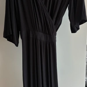 Svart omlottklänning från KappAhl - Svart långklänning från KappAhl med omlottmodell och knytband i midjan. Klänningen har korta, vida ärmar och ett mjukt, lätt material som faller snyggt. Perfekt för dig som gillar en klassisk och stilren look.