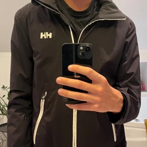 Svart vindjacka från Helly Hansen - Svart vindjacka från Helly Hansen med vit dragkedja och logga på bröstet. Jackan har huva, två snedställda fickor med dragkedja och en sportig look. Perfekt för blåsiga dagar och enkel att matcha med det mesta.