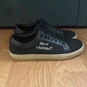 Svarta Saint Laurent sneakers - Svarta sneakers från Saint Laurent med vit text på sidan och klassisk vit sula. Skorna är jätte fräscha och snygga till allt säljer pågrund av att dem är för småa skorna är helt nya men har testat dem en dag skriv gärna vid frågor 😝