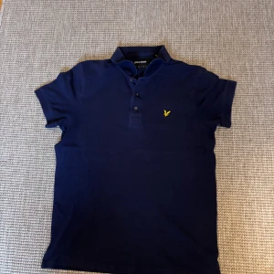 Mörkblå pikétröja från Lyle & Scott - Mörkblå pikétröja från Lyle & Scott med klassisk krage, tre knappar och den ikoniska gula fågeln broderad på bröstet. Tröjan är i bomull och har korta ärmar, perfekt för en clean och stilren look.