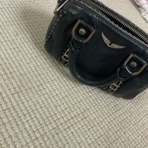 Svart handväska Zadig & Voltaire skinn - Cool svart handväska från Zadig & Voltaire i äkta skinn med silvriga nitar och detaljer. Väskan har dubbla handtag, dragkedja upptill och ikonisk vinge framtill. Perfekt storlek för det viktigaste och ger en edgy vibe till din outfit.