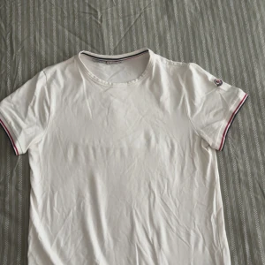 Vit t-shirt från Moncler med logga - Vit t-shirt från Moncler med klassisk rund hals och korta ärmar. Ärmsluten har snygga ränder i rött och blått samt en liten Moncler-logga på vänster ärm. Tillverkad i mjuk bomull för en skön känsla.
