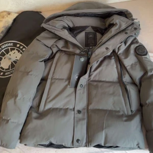 Grå Canada Goose wyndham  - Säljer en grå dunjacka från Canada Goose med klassisk patch på ärmen. Jackan har huva, dragkedja och knappar framtill samt stora fickor med dragkedja. Perfekt för kalla dagar och riktigt snygg till streetwear.