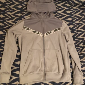 Grå Nike hoodie (147-158 barnstorlek) - Nike hoodie i ljusgrått med hel dragkedja och huva. Snygg detalj med vit Nike-logga och mönstrad rand över bröstet. Tillverkad i Dri-FIT-material som andas, perfekt för träning eller chill. Fickor framtill och ribbade muddar för extra komfort. Storlek 147-158 barn. Kan förekomma katthår.