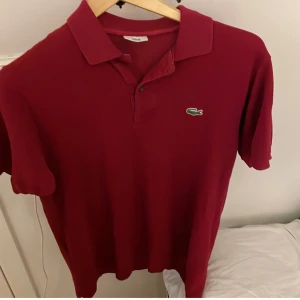 Röd pikétröja från Lacoste - Klassisk röd pikétröja från Lacoste med den ikoniska gröna krokodilloggan på bröstet. Tröjan har krage, korta ärmar och knäppning framtill. Tillverkad i bomull för en skön och luftig känsla. Perfekt för en clean och sportig stil.