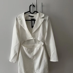 Vit cut out blazerklänning Boohoo - Supersnygg vit kort blazerklänning från Boohoo med cut out-detalj i midjan och klassisk krage. Klänningen har lång ärm och stängs med en knapp framtill. Perfekt för dig som vill sticka ut med en trendig look. Klänningen är oanvänd.