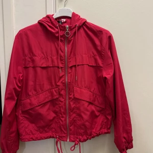 Röd vindjacka från H&M Divided XS - Snygg röd vindjacka från H&M Divided i storlek XS. Jackan har huva, dragkedja med rund metallring, snörning i midjan och stora fickor framtill. Tillverkad i lätt polyester med meshfoder upptill för extra komfort. Perfekt för vår och höst.