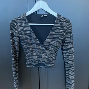 Glittrig croppad topp från H&M Divided - Säljer en svart croppad topp från H&M Divided i storlek XS. Toppen har glittrigt zebramönster, lång ärm och djup v-ringning. 
