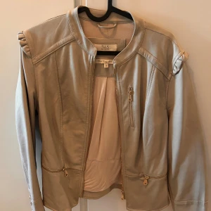 Beige skinnjacka från 365, strl 38 - Snygg beige skinnjacka från 365 med guldfärgade dragkedjor och coola rynkade detaljer på axlarna. Jackan har rundad krage, flera fickor med dragkedja och en stilren, figurnära passform. 