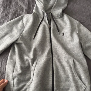 Grå hoodie från Polo Ralph Lauren - Snygg grå hoodie från Polo Ralph Lauren med svart logga på bröstet. Tröjan har dragkedja, huva med svarta snören och två stora fickor framtill. Perfekt för chill dagar eller när du vill ha en clean och stilren look.