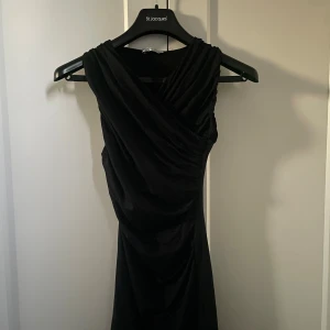 Svart omlottklänning med drapering - Snygg svart klänning med detalj kring bröstet och draperad effekt över byst och midja. Ärmlös, kort och figurnära passform. 