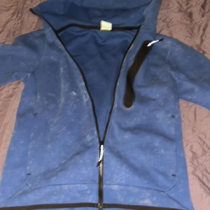 Blå Nike ziphoodie med huva - Säljer en blå hoodie med dragkedja från Nike med huva och svart dragkedja som går snett över bröstet. Jackan har en bröstficka med dragkedja och är långärmad. Perfekt för dig som gillar sportig stil och vill ha något bekvämt och snyggt.