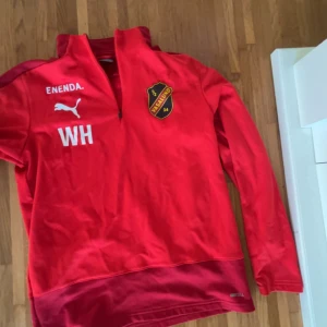 Röd Puma Vasalund träningströja - Röd långärmad träningströja från Puma med Vasalund-emblem på bröstet och initialerna WH. Tröjan har halv dragkedja framtill och är perfekt för fotbollsträning. Snygg sportig look med tryckta detaljer och hög krage.