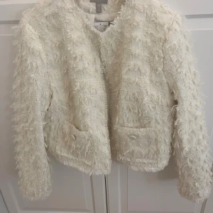 Vit fransig kavaj från H&M, storlek L - Säljer en vit kavaj från H&M med fransiga detaljer och två fickor framtill. Jackan har en strukturerad, vävd yta och knappar på fickorna. Aldrig använd, med prislapp!