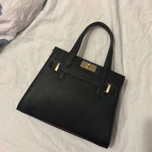 Svart handväska från Bershka - Stilren svart handväska från Bershka med guldfärgade metalldetaljer och strukturmönstrad yta. Väskan har både handtag och avtagbar axelrem, samt snyggt lås framtill. Perfekt för dig som gillar klassisk och trendig design.