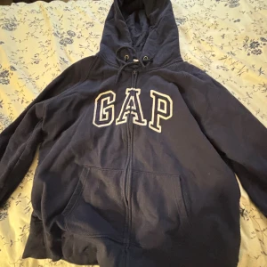Mörkblå GAP hoodie med dragkedja - Klassisk mörkblå hoodie från GAP med stor vit broderad logga på bröstet. Tröjan har huva med snörning, dragkedja framtill och en stor känguruficka. Perfekt för chill dagar eller när du vill ha en avslappnad look.