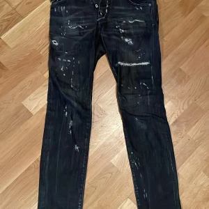 Svarta Dsquared2 jeans - Säljer ett par svarta jeans från Dsquared2 med coola slitningar och färgstänk för en edgy look. Jeansen har klassisk femficksdesign, smal passform och är tillverkade i stretchig bomull. Perfekta för dig som vill sticka ut med en streetinspirerad stil.