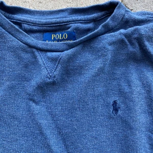 Blå tröja från Polo Ralph Lauren - Klassisk blå tröja från Polo Ralph Lauren med rund halsringning och diskret broderad logga på bröstet. Tröjan har långa ärmar och ribbade muddar. Perfekt för dig som gillar enkel och stilren design. Aningen tvättblekt. Behöver strykas. Bomull. Passar XS-S  eller 14-16 år
