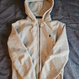 Grå hoodie från Polo Ralph Lauren - Säljer en ljusgrå hoodie från Polo Ralph Lauren med dragkedja och huva. Tröjan har snörning i huvan, klassisk logga broderad på bröstet och är tillverkad i mjuk bomullsmix. Perfekt för chill dagar eller när du vill ha en clean look.