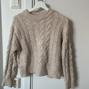 Beige stickad tröja från Gina Tricot - Mysig beige stickad tröja från Gina Tricot med grovt kabelstickat mönster. Tröjan har ribbade muddar vid hals, ärmslut och nederkant samt lång ärm. Perfekt för kyliga dagar och enkel att styla med jeans eller kjol.