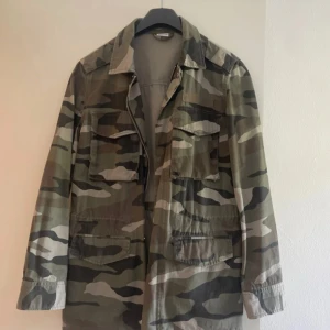 Camouflage parkajacka med snörning - Säljer en grön och beige camouflage parkajacka med dragkedja och stora fickor framtill. Jackan har snörning i midjan för justerbar passform och klassisk krage. Perfekt för dig som gillar militärinspirerad stil.