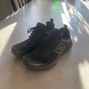 Svarta Nike Presto sneakers - Svarta Nike Presto sneakers med snygg, strömlinjeformad design och platt sula. Skorna har snörning och en flexibel ovandel i syntetmaterial som ger en skön passform. Sidorna har en cool plastdetalj med Nike-logga och rund tå. Perfekta för dig som gillar sportig stil.