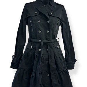 Svart trenchcoat i lolita-stil - Svart trenchcoat med dubbelknäppning, markerad midja med bälte och volangliknande nederdel. Jackan har långa ärmar, axelklaffar, dragkedjefickor och dekorativa knappar. Snygg och edgy look som lyfter din outfit.