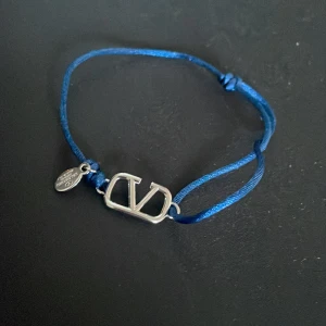 Blå Valentino armband med silverdetalj - Snyggt armband från Valentino med blå justerbar tråd och en silverfärgad logodetalj i metall. Perfekt accessoar för att ge din outfit en lyxig touch. Liten bricka med Valentino-gravyr hänger bredvid logon.