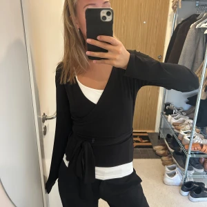 Svart omlottopp med knytband - Svart omlottopp med långa ärmar och knytband i midjan. Toppen har en v-ringad skärning och är i mjukt, stretchigt material. Perfekt att styla med ett linne under för en snygg lager-på-lager-look.