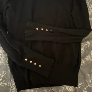 Svart stickad tröja från Zara med knappar - Svart stickad tröja från Zara i storlek S. Tröjan har långa ärmar med snygga guldfärgade knappar vid ärmsluten och rund halsringning. Perfekt för att lyfta din outfit med en stilren touch.