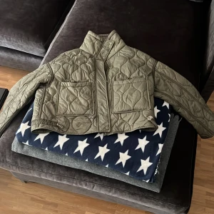 Olivgrön quiltad pufferjacka - Säljer en olivgrön quiltad pufferjacka med två stora fickor framtill och hög krage. Jackan har en croppad passform och stängs med dragkedja. Snygg vågig quiltning över hela jackan ger en unik look. Perfekt för dig som vill ha en trendig och bekväm jacka.