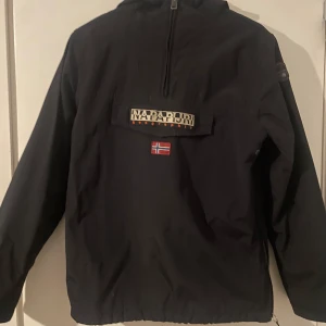  Napapijri jacka - Svart vindjacka från Napapijri med halv dragkedja och stor logga samt norsk flagga på bröstet. Jackan har huva, fleecefoder på insidan och praktisk magficka med lock. Perfekt för kyliga dagar. Tar emot byten.