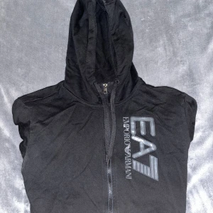 Svart EA7 kofta  - Svart kofta från Emporio Armani EA7 med stor logga på bröstet. Tröjan har huva med snörning, hel dragkedja och fickor framtill. Perfekt för dig som gillar stilrena och sportiga plagg.