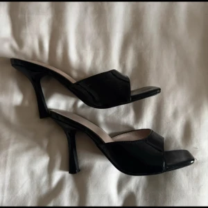 Svarta klackade mules med öppen tå - Snygga svarta mules med hög klack och öppen tå. Skorna har en glansig finish och fyrkantig tå, vilket ger en trendig look. Perfekta att styla till kjol eller jeans för en chic vibe.