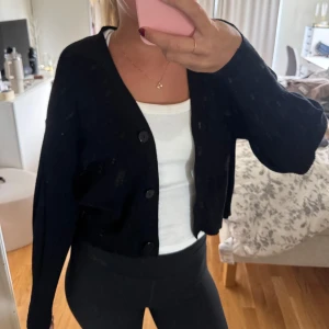 Svart croppad kofta med knappar - Svart croppad kofta med tre stora knappar framtill och v-ringad hals. Koftan har lång ärm och är stickad i ett mjukt material som känns skönt mot huden. 