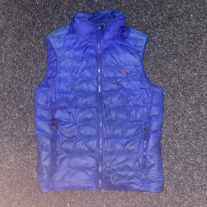 Blå dunväst från Polo Ralph Lauren - Blå dunväst från Polo Ralph Lauren med quiltad design och hög krage. Västen har dragkedja framtill, två fickor med dragkedja och en liten röd logga broderad på bröstet. Perfekt för lager-på-lager och chill stil.