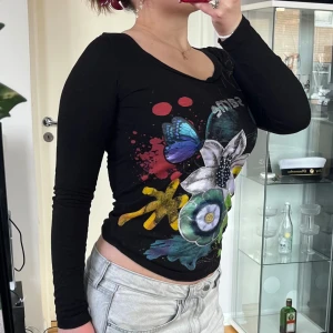Svart långärmad topp med blommor XS - Svart långärmad topp från Desigual i bomull med färgglatt blommönster, fjäril och text framtill. Snyggt snörningsband med röd tofs i nacken. Tight passform och rund halsringning. Perfekt för dig som gillar unika prints och detaljer. Axelbredd: 35cm. Längd: 60cm. 