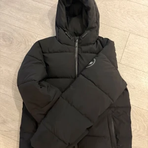 Svart pufferjacka Supply & Demand - Svart pufferjacka från Supply & Demand med huva och dragkedja framtill. Jackan har stora vadderade paneler, två fickor med dragkedja och tryckt SUPPLY-logga på ryggen. Perfekt för kalla dagar och har en clean streetstil. Storlek 13-15 år men passar som S i herr. Köpt från JD Sports för va 1199kr, varan är i mycket bra skick. 