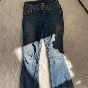 True Religion blå bootcut jeans - Priset går att diskutera!   Säljer ett par klassiska blå jeans från True Religion med bootcut passform. Jeansen har slitna detaljer på benen, ikoniska bakfickor med broderi och silverfärgade knappar. Tillverkade i USA i mjukt denimtyg med snygga kontrastsömmar.
