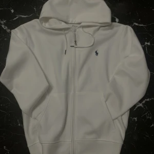 Vit hoodie från Polo Ralph Lauren med dragkedja  - Vit hoodie från Polo Ralph Lauren med dragkedja och huva. Klassisk design med broderad svart polo logga på bröstet. Perfekt för en clean och stilren look.