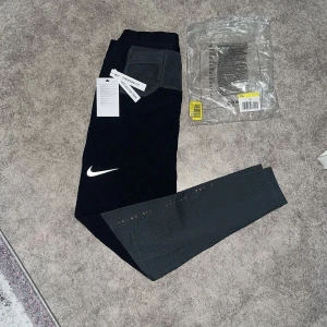 Svarta och grå Nike träningsbyxor - Nike träningsbyxor i svart och grått med reflekterande detaljer längs benen och klassisk vit Swoosh-logga. Byxorna har elastisk midja med snörning, praktiska fickor och ventilerande meshpaneler på underbenen. Perfekta för dig som vill ha stil och funktion på gymmet.