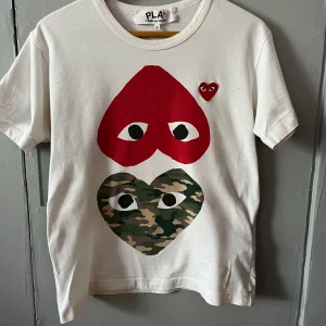 Vit Comme des Garçons Play t-shirt S - Vit t-shirt från Comme des Garçons Play med ikoniska hjärttryck framtill, ett i rött och ett i camouflage. Klassisk passform, rund hals och korta ärmar. Tillverkad i 100% bomull för en skön känsla. Perfekt statement-plagg för dig som gillar streetstyle. 