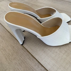 Vita klackade sandaler med fyrkantig tå - Snygga vita sandaler med hög klack och fyrkantig tå. Skorna har en bred rem i croco-mönstrat syntetmaterial och öppen häl. Perfekta för dig som vill sticka ut med en trendig och stilren look.