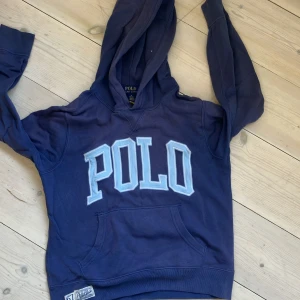 Blå hoodie från Polo Ralph Lauren, strl 10-12 - Blå hoodie från Polo Ralph Lauren med stor ljusblå POLO-text framtill. Fint skick 