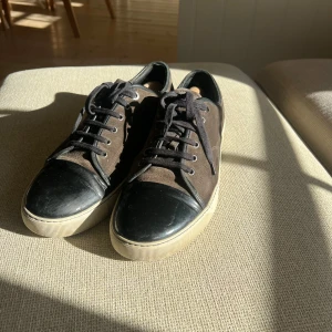 Bruna lanvins - Snygga sneakers i brun mocka med svart tåparti i skinn och svarta snören. Skorna har en vit sula och klassisk rund tå. Perfekta för dig som gillar en stilren men ändå unik look. Skick 7/10