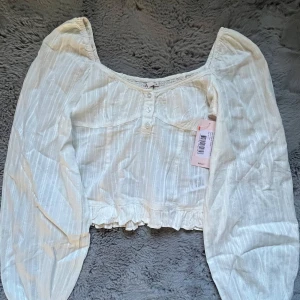 Vit blus med puffärm från Nelly - Supersöt vit blus från Nelly med puffiga långa ärmar, knappar framtill och diskret randigt mönster. Blusen har croppad passform, volangkant nertill och lätt v-ringad hals. Helt ny med prislapp och aldrig använd.